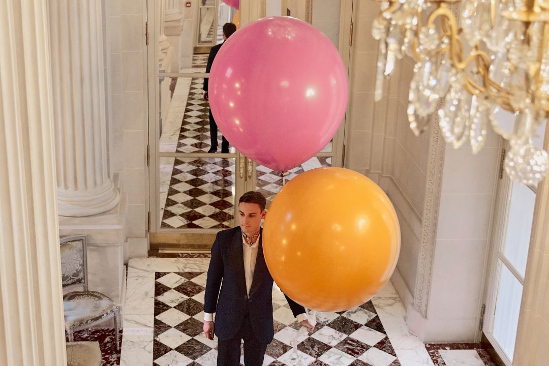 A man in a suit stands in a hallway with large  pink and orange balloons.